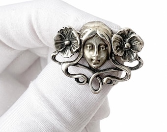 May include: A silver-toned brooch featuring a stylized female face with flowing hair, flanked by two detailed flower designs. The brooch has an Art Nouveau style with swirling lines and floral elements. The piece is held against a white background.