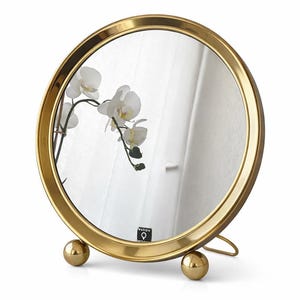 Könnte beinhalten: Ein runder, goldfarbener Schminkspiegel mit Messingrahmen und -fuß. Der Spiegel reflektiert eine weiße Orchidee und eine hellfarbige Wand. Der Fuß hat zwei goldene Kugeln und eine gebogene Stütze. Der Spiegel hat unten den Text "MASSI".