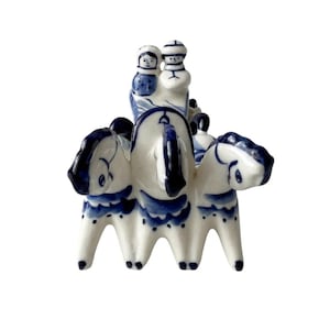 May include: A white ceramic figurine featuring three horses pulling a carriage with two figures seated inside. The piece is decorated with hand-painted blue patterns, including detailed manes, facial features, and ornate trim on the horses and carriage.