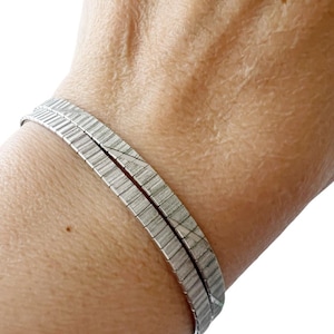 Op de afbeelding: Twee zilverkleurige armbanden met een getextureerd, rechthoekig ontwerp. De armbanden worden om een pols gedragen. De afbeelding is van dichtbij genomen. De armbanden lijken van metaal te zijn en hebben een flexibele constructie.