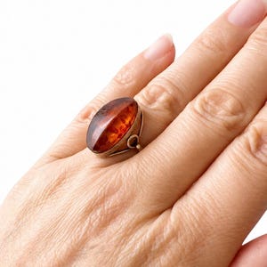 May include: A silver ring featuring a large, oval amber stone. The amber has a rich, warm brown color with internal patterns. The ring is set on a hand, showcasing the jewelry's design and size. The ring's setting has a decorative loop.