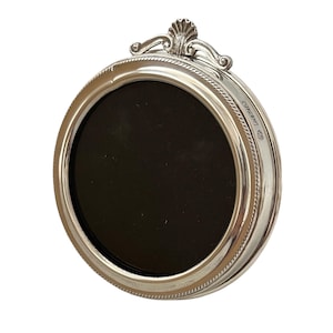 May include: A circular silver picture frame with a polished metallic finish and a rope-style border. The top features a decorative shell-shaped crest. The frame contains a solid black circular insert and displays small engraved hallmarks on the side.