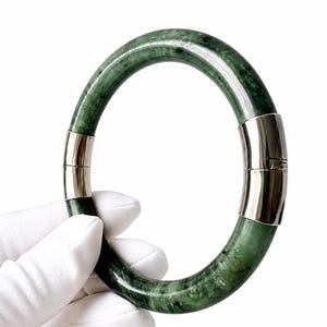 Könnte beinhalten: Ein grünes Jade-Armreif-Armband mit silberfarbenen Metallakzenten. Das Armband hat eine solide, kreisförmige Form mit poliertem Finish. Die Metallakzente befinden sich an den Seiten des Armbands, und der Jade hat ein marmoriertes Aussehen.