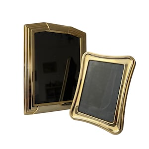 May include: Two gold-coloured picture frames. One is rectangular with a stepped design, and the other is a smaller, curved frame. Both have black interiors, contrasting with the shiny gold finish.