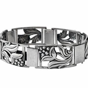 Puede incluir: Pulsera plateada con un patrón repetitivo de eslabones rectangulares y motivos decorativos florales y de racimos de uvas. La pulsera es de metal y tiene un cierre de broche. Joyería sobre fondo blanco.