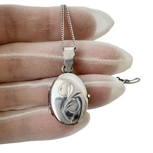 Può includere: Ciondolo a forma di medaglione ovale in argento con un ornato motivo floreale. Il medaglione è appeso a una delicata catena d'argento. Il ciondolo è lungo circa 2,5 cm e ha un'apertura a cerniera.