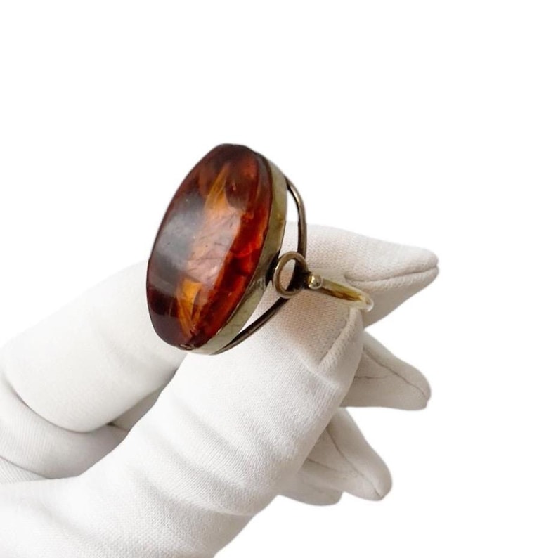 May include: An oval amber-colored gemstone ring with a gold-colored band. The amber stone is set in a silver-colored metal frame. The ring is held by a gloved hand against a white background. The amber stone has a rich, warm color.