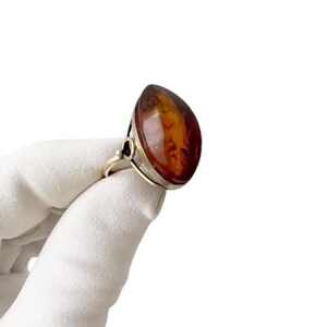 May include: A silver ring featuring a large, teardrop-shaped amber stone. The amber has a warm, reddish-brown hue with internal patterns. The ring's band has a simple, elegant design.