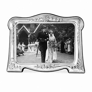May include: A silver picture frame with an elaborate design, containing a black and white wedding photograph. The frame features a curved top and floral embellishments. The photograph depicts a bride and groom walking along a path.