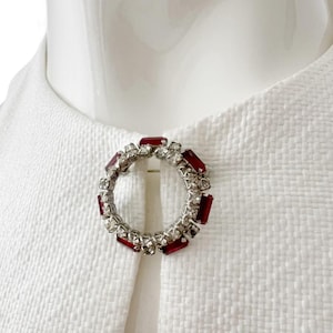 Puede incluir: Broche circular plateado con piedras rectangulares rojas y transparentes alternas. El broche está prendido a una tela blanca texturizada, creando un contraste. Un accesorio clásico.