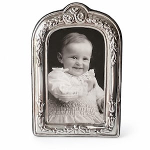 May include: An arched silver picture frame featuring embossed floral and leaf motifs along the border. The frame contains a black and white photograph of a smiling infant wearing a smocked dress with ruffled sleeves, captured in a portrait orientation.
