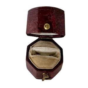 May include: An open, antique burgundy ring box with a gold clasp and button closure. The interior is lined with a cream-colored velvet material. The box is octagonal in shape.