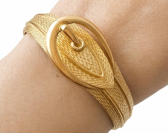 May include: A gold-coloured bracelet with a buckle design. The bracelet has a textured band and a large, oval-shaped buckle. The buckle has a textured surface and a curved bar. The jewellery is worn on a wrist.