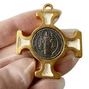 Puede incluir: Un colgante de cruz dorado con detalles de esmalte blanco. El centro presenta un medallón plateado con una figura religiosa y texto. El colgante tiene un lazo en la parte superior para colgar.