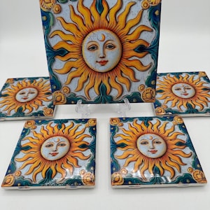 May include: A set of ceramic tiles featuring a sun design. The sun has a face with a crescent moon above it, surrounded by yellow rays. The tiles have a blue and green border. The set includes one larger tile and four smaller tiles.