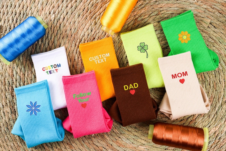 Op de afbeelding: Assortiment van kleurrijke sokken met geborduurde ontwerpen en tekst, waaronder "Custom Text", "Follow Your", "Dad", "Mom" en een klavertjevier en een bloem.