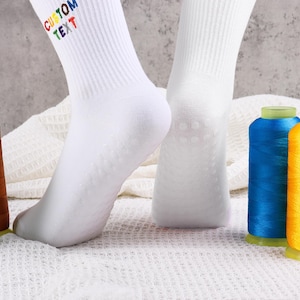 Op de afbeelding: Witte sokken met de tekst "CUSTOM TEXT" in regenboogkleuren op de bovenkant van de sok. De sokken liggen op een wit, pluche oppervlak met spoelen van groene, bruine, blauwe en gele draad.