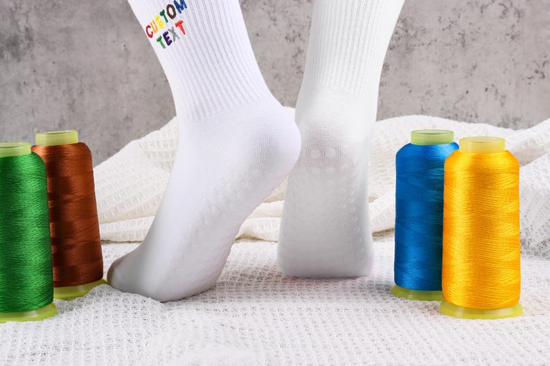 Op de afbeelding: Witte sokken met de tekst "CUSTOM TEXT" in regenboogkleuren op de bovenkant van de sok. De sokken liggen op een wit, pluche oppervlak met spoelen van groene, bruine, blauwe en gele draad.