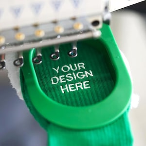 Op de afbeelding: Een close-up van een groene stof die wordt geborduurd met wit garen. De borduurwerktekst is "YOUR DESIGN HERE".