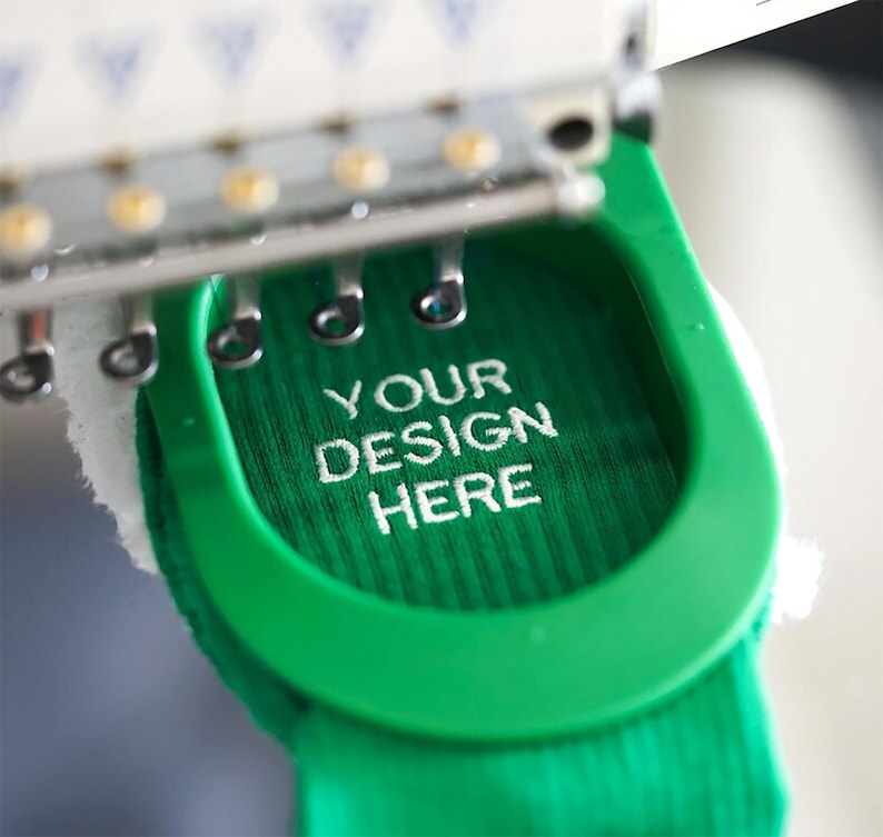 Op de afbeelding: Een close-up van een groene stof die wordt geborduurd met wit garen. De borduurwerktekst is "YOUR DESIGN HERE".