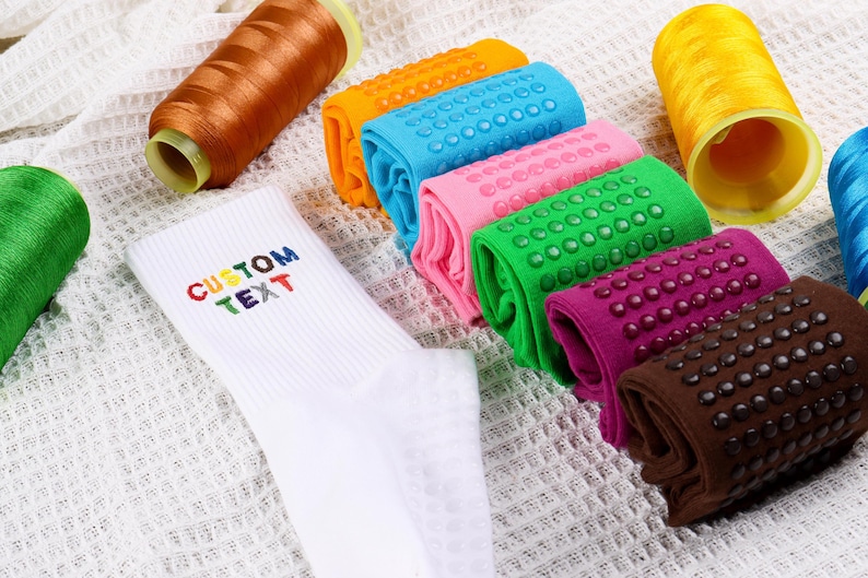 Op de afbeelding: Een witte sok met de tekst "CUSTOM TEXT" geborduurd in regenboogkleuren. De sok ligt op een wit oppervlak met spoelen garen in verschillende kleuren, waaronder oranje, geel, groen en roze.