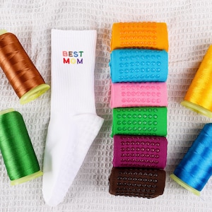 Op de afbeelding: Witte sokken met de tekst "BEST MOM" geborduurd in regenboogkleuren. De sokken worden getoond met spoelen garen in verschillende kleuren, waaronder oranje, groen, roze, blauw en bruin.