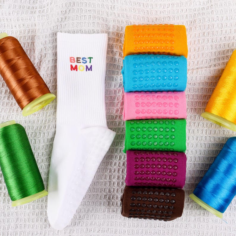 Op de afbeelding: Witte sokken met de tekst "BEST MOM" geborduurd in regenboogkleuren. De sokken worden getoond met spoelen garen in verschillende kleuren, waaronder oranje, groen, roze, blauw en bruin.