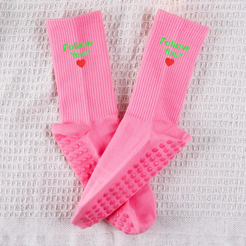 Op de afbeelding: Een paar roze sokken met gripdots op de onderkant. De sokken hebben de tekst "Follow Your" in groen met een klein rood hartje.