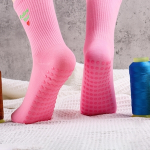 Op de afbeelding: Een paar roze sokken met een groene en witte "Follow Your Heart" afbeelding en een rood hart. De sokken hebben een textuur grip aan de onderkant. Drie spoelen garen zijn op de achtergrond: groen, bruin en blauw.