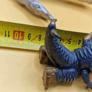 Puede incluir: Primer plano de una criatura de juguete azul y dorada con una cinta m&eacute;trica. La criatura tiene un dise&ntilde;o detallado con un cuerpo azul oscuro y detalles dorados. La cinta m&eacute;trica muestra medidas en cent&iacute;metros.