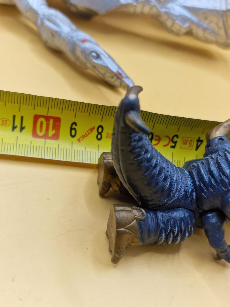 Puede incluir: Primer plano de una criatura de juguete azul y dorada con una cinta m&eacute;trica. La criatura tiene un dise&ntilde;o detallado con un cuerpo azul oscuro y detalles dorados. La cinta m&eacute;trica muestra medidas en cent&iacute;metros.