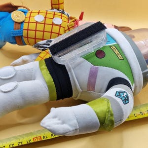 May include: Two plush toy figures: Woody in a yellow plaid shirt and blue pants, and Buzz Lightyear in a green and white spacesuit. Buzz has a clear helmet and the word "Lightyear" on his arm. A measuring tape is in the foreground.