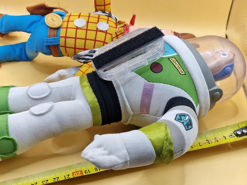 May include: Two plush toy figures: Woody in a yellow plaid shirt and blue pants, and Buzz Lightyear in a green and white spacesuit. Buzz has a clear helmet and the word "Lightyear" on his arm. A measuring tape is in the foreground.