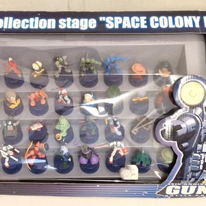 May include: A boxed set of Pepsi Twist bottle cap figures. The box reads "The collection stage SPACE COLONY MODEL" and features a clear window displaying the collection of small, colorful figures. The box also features Gundam artwork.