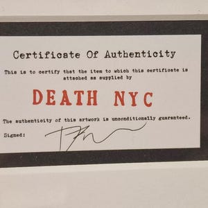 May include: A black and white certificate of authenticity with the text "Certificate of Authenticity" and "DEATH NYC" in red. The text "This is to certify that the item to which this certificate is attached as supplied by" and "The authenticity of this artwork is unconditionally guaranteed." is printed in black. The certificate is signed with a black ink signature.