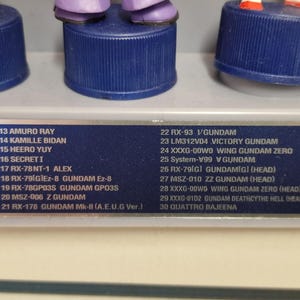 May include: A close-up of a display of Gundam figures, each mounted on a blue bottle cap base. The figures are in various colors, with a list of names and model numbers visible below. The text includes names like Amuro Ray and Kamille Bidan.