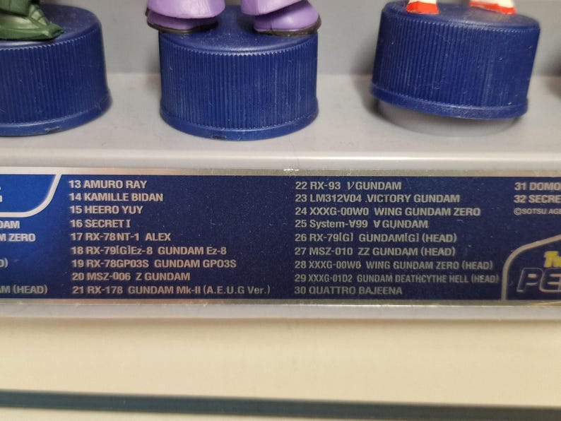 May include: A close-up of a display of Gundam figures, each mounted on a blue bottle cap base. The figures are in various colors, with a list of names and model numbers visible below. The text includes names like Amuro Ray and Kamille Bidan.