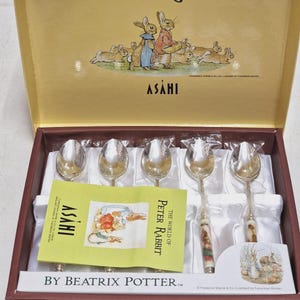 May include: A boxed set of Peter Rabbit spoons. The gold box features illustrations of Peter Rabbit characters. The silver spoons have decorative handles. A small booklet with illustrations and the text "The World of Peter Rabbit" is also included in the box.