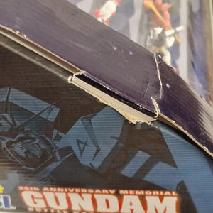 May include: Close-up of a partially visible box for a Gundam collectible. The box is primarily dark blue with a graphic of a Gundam robot and the text "25th Anniversary Memorial Gundam Bottle Cap". The box has some damage.