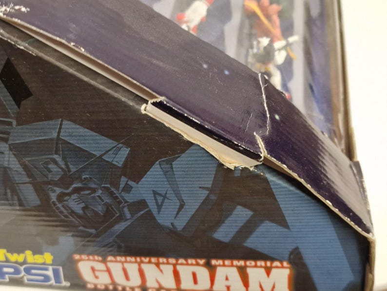 May include: Close-up of a partially visible box for a Gundam collectible. The box is primarily dark blue with a graphic of a Gundam robot and the text "25th Anniversary Memorial Gundam Bottle Cap". The box has some damage.