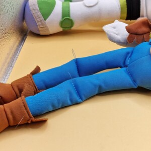 May include: Close-up of a plush toy featuring Woody's legs in blue jeans and brown boots. A large button and belt buckle detail the waist. Buzz Lightyear's arm is visible in the background, with green and white accents.
