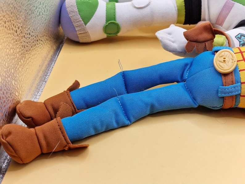 May include: Close-up of a plush toy featuring Woody's legs in blue jeans and brown boots. A large button and belt buckle detail the waist. Buzz Lightyear's arm is visible in the background, with green and white accents.