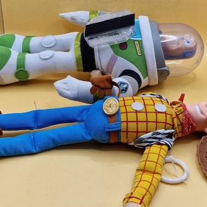 May include: Two plush toys from the Toy Story franchise. One is Buzz Lightyear, in a green, white, and black spacesuit with a clear helmet. The other is Woody, a cowboy doll with a yellow plaid shirt, blue pants, and a brown cowboy hat.