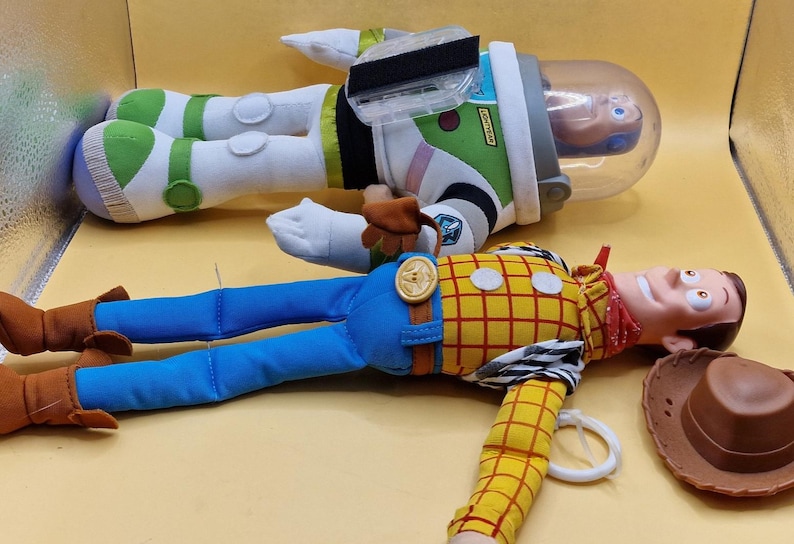 May include: Two plush toys from the Toy Story franchise. One is Buzz Lightyear, in a green, white, and black spacesuit with a clear helmet. The other is Woody, a cowboy doll with a yellow plaid shirt, blue pants, and a brown cowboy hat.
