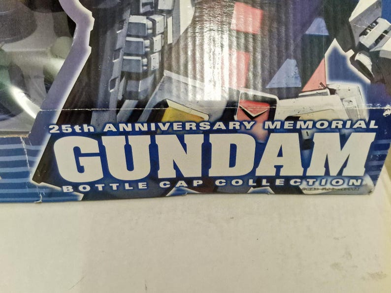 May include: Close-up of a box with the text "25th Anniversary Memorial Gundam Bottle Cap Collection" in white and blue. The box has a dark blue background with images of a Gundam robot.