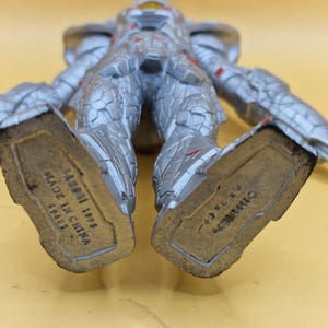Puede incluir: Figura de acci&oacute;n plateada y roja con intrincados detalles de armadura. La figura est&aacute; tumbada boca arriba, con la parte inferior de los pies visible, mostrando el texto "BANDAI 1996 MADE IN CHINA".