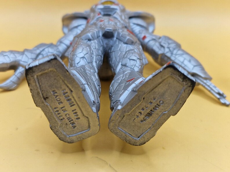 Puede incluir: Figura de acci&oacute;n plateada y roja con intrincados detalles de armadura. La figura est&aacute; tumbada boca arriba, con la parte inferior de los pies visible, mostrando el texto "BANDAI 1996 MADE IN CHINA".