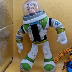May include: A plush Buzz Lightyear doll from the Toy Story franchise. The doll is wearing a white and green spacesuit with a clear helmet. The word "LIGHTYEAR" is printed on the chest. The doll has a smiling face and blue eyes.