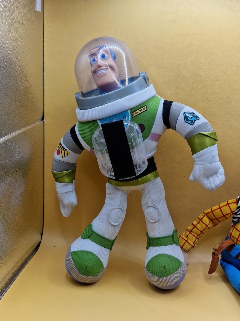 May include: A plush Buzz Lightyear doll from the Toy Story franchise. The doll is wearing a white and green spacesuit with a clear helmet. The word "LIGHTYEAR" is printed on the chest. The doll has a smiling face and blue eyes.