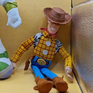May include: A Woody doll from the Toy Story franchise. The doll is wearing a brown cowboy hat, a yellow plaid shirt, blue pants, and brown boots. A red bandana is around the neck. The doll has a friendly expression.
